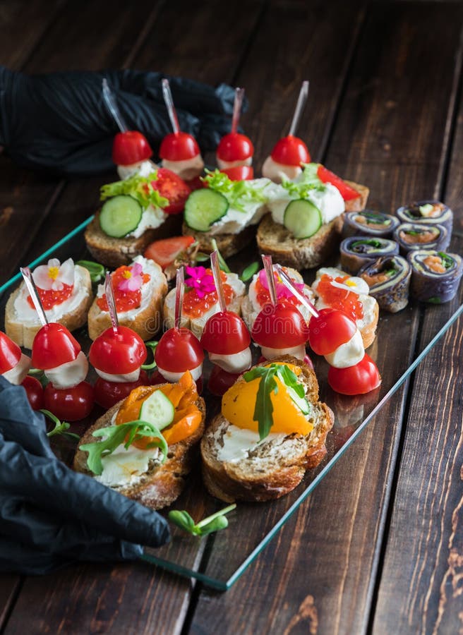 Två händer i svarta handskar som rymmer ett mellanmålmagasin på träig bakgrund: rullar canapes, rullar, välfyllda tomater, smörgå arkivbild Två händer i svarta handskar som rymmer ett mellanmålmagasin på träig bakgrund: rullar canapes, rullar, välfyllda tomater, smörgå arkivbild