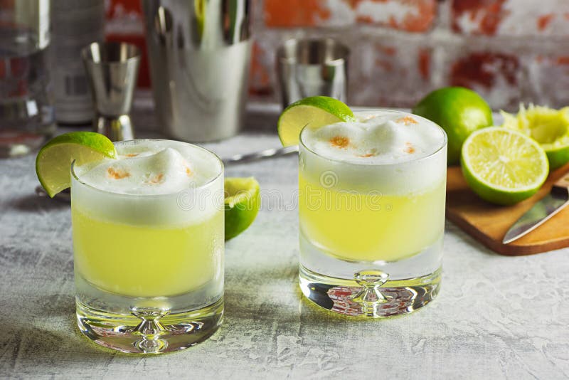 Two rocks glasses filled with pisco sours and garnished with lime wedges on a bar top. This cocktail is made from Peruvian pisco liquor, lime juice, simple syrup, and egg white shaken together and topped with a couple dashes of bitters. The tools and ingredients used to make the drinks are in the background. Two rocks glasses filled with pisco sours and garnished with lime wedges on a bar top. This cocktail is made from Peruvian pisco liquor, lime juice, simple syrup, and egg white shaken together and topped with a couple dashes of bitters. The tools and ingredients used to make the drinks are in the background.