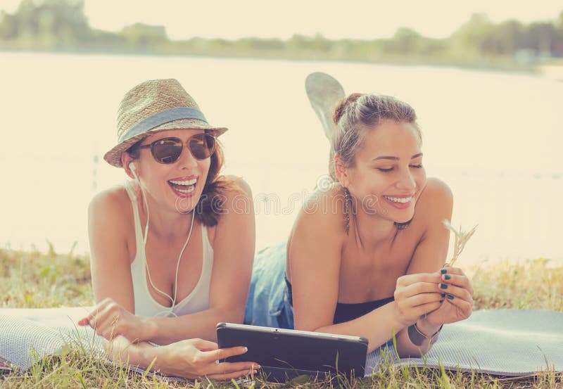 Two funny happy young women. Friends laughing browsing watching social media videos listen to music on smart pad computer, laying outdoors on green meadow on a sunny summer day. Two funny happy young women. Friends laughing browsing watching social media videos listen to music on smart pad computer, laying outdoors on green meadow on a sunny summer day