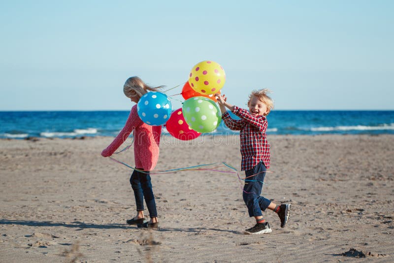 Två roliga vita Caucasian barnungar med den färgrika gruppen av ballonger som spelar att köra på stranden på solnedgång, höstsomm arkivfoto Två roliga vita Caucasian barnungar med den färgrika gruppen av ballonger som spelar att köra på stranden på solnedgång, höstsomm arkivfoto