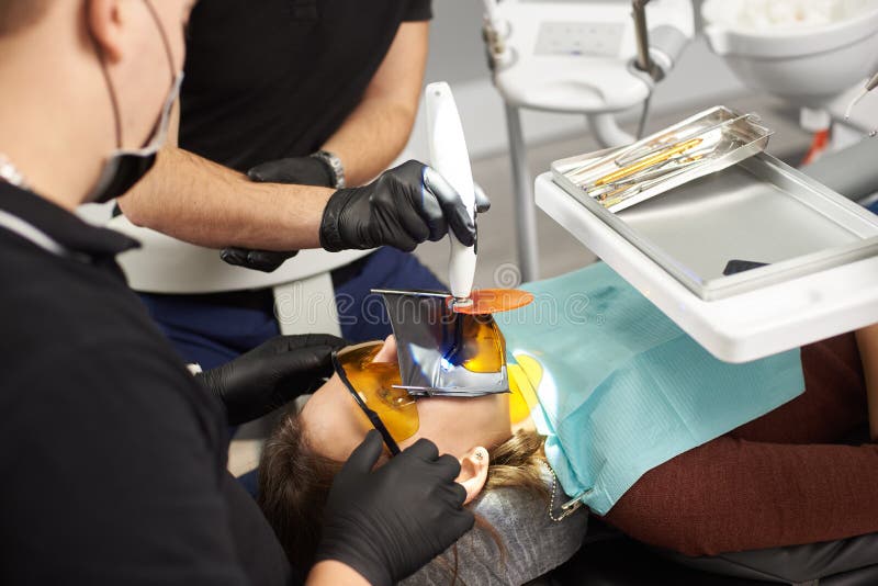 Två tandläkare i svarta dräkter som fixar tänderna av en patient med en kofferdam på framsidan royaltyfri fotografi Två tandläkare i svarta dräkter som fixar tänderna av en patient med en kofferdam på framsidan royaltyfri fotografi