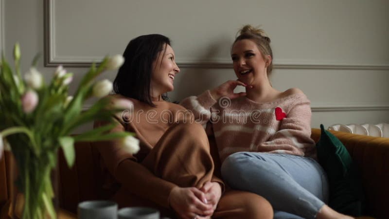 Twee gelukkige jonge vrouwen vrienden op sofa bij bouquet van witte tulpen bloemen thuis terwijl ze praten over roddelen.