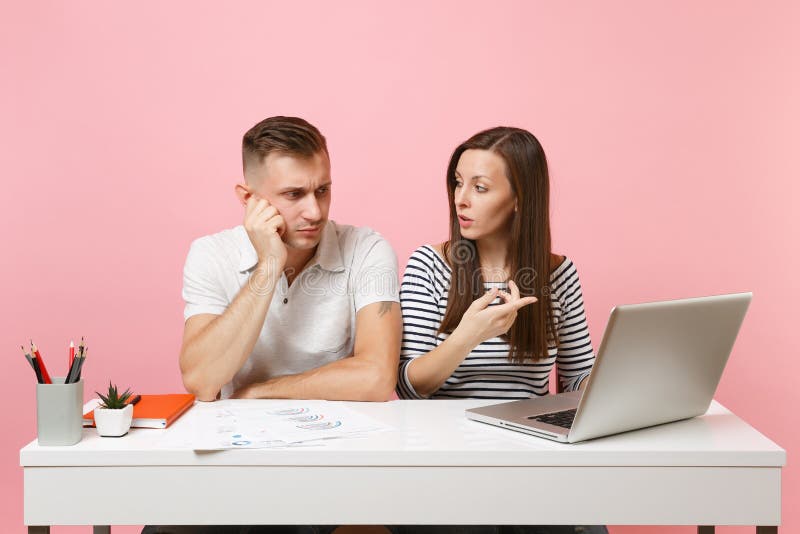 Twee glimlachende bedrijfsvrouwenman collega's zitten het werk bij wit bureau met eigentijdse laptop op pastelkleur roze achtergr stock fotografie Twee glimlachende bedrijfsvrouwenman collega's zitten het werk bij wit bureau met eigentijdse laptop op pastelkleur roze achtergr stock fotografie