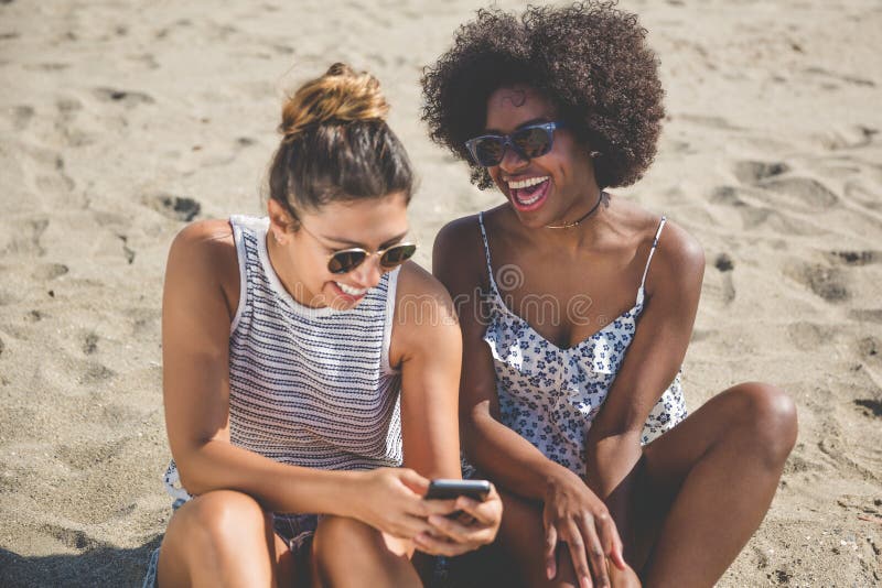 Twee Meisjes Op Strand Die Het Mobiele Lachen Gebruiken Stock Foto