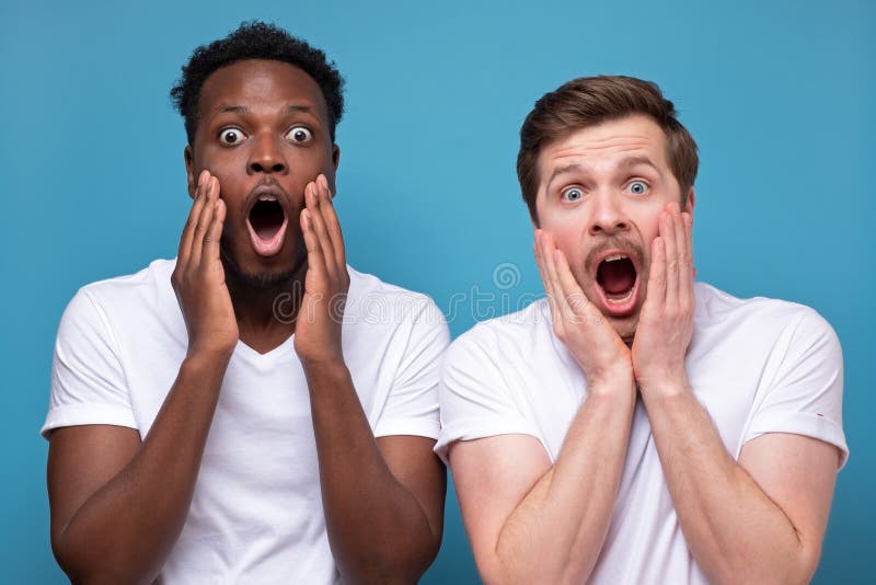 Two african and caucasian men looking with shocked expression, with mouth wide open, holding hands on face, posing isolated against blue background