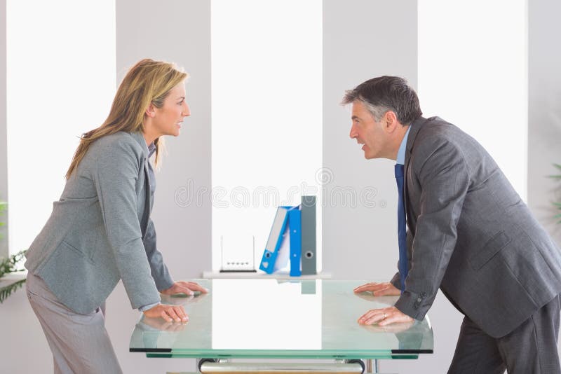 Two angry businesspeople standing arguing on each side of a desk at office. Two angry businesspeople standing arguing on each side of a desk at office