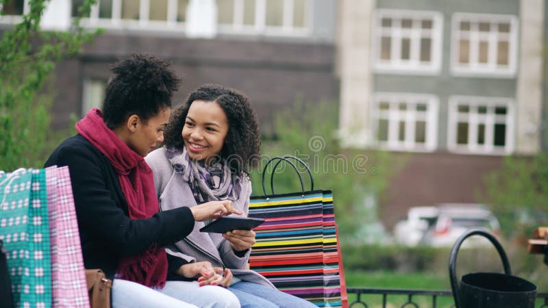 Two attractive mixed race women with shopping bags sitting on bench and using tablet computer for internet surfing stock image Two attractive mixed race women with shopping bags sitting on bench and using tablet computer for internet surfing stock image