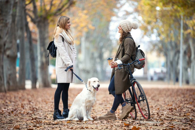 Two beautiful friends talking while walking with their dog and bike in the park in autumn