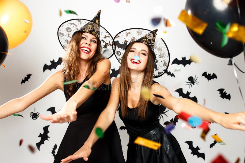 Two beautiful girls in black dresses and witch hats have fun with balloons and confetti on the background of the wall with bats .