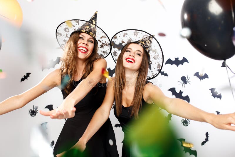 Two beautiful girls in black dresses and witch hats have fun with balloons and confetti on the background of the wall with bats .