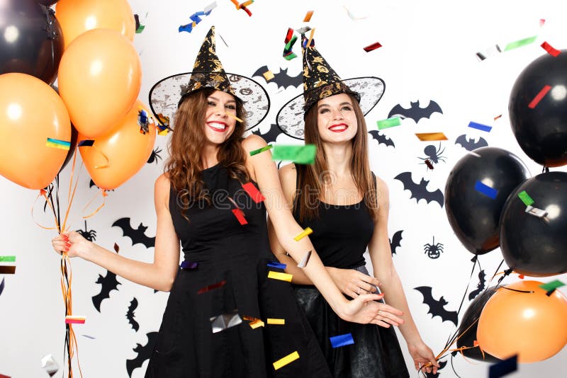 Two beautiful girls in black dresses and witch hats have fun with black and orange balloons and confetti.Halloween party .