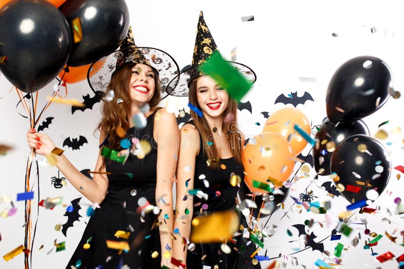 Two beautiful girls in black dresses and witch hats have fun with black and orange balloons and confetti. Halloween party .