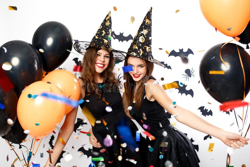 Two beautiful girls in black dresses and witch hats have fun with black and orange balloons and confetti. Halloween party .