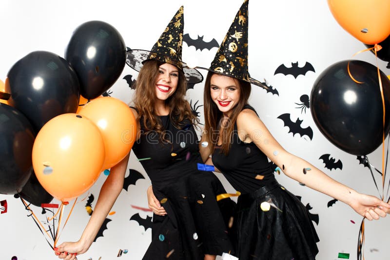 Two beautiful girls in black dresses and witch hats have fun with black and orange balloons and confetti. Halloween party .