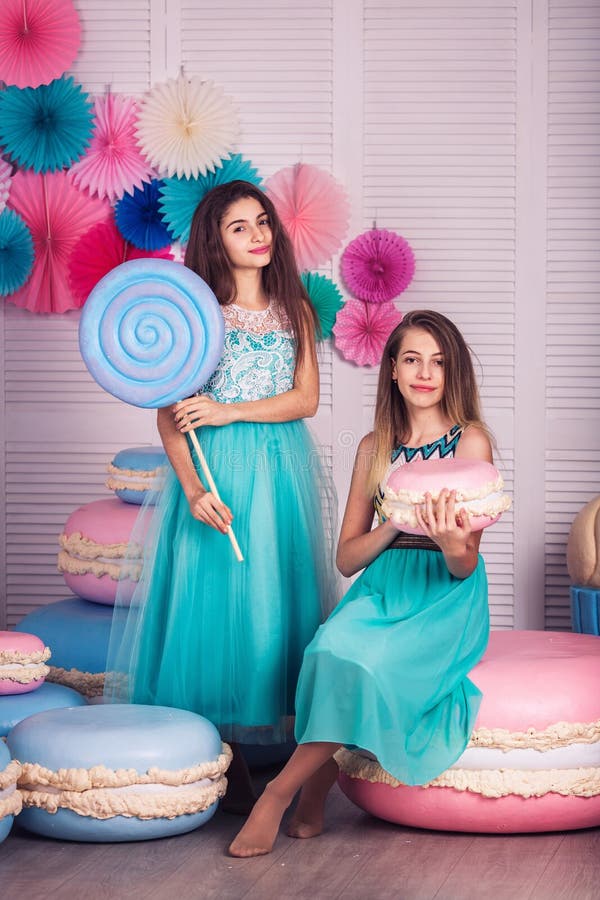 Two beautiful girls in blue dresses hold in their hands huge candy and macaroon in studio with decor of macaroons.