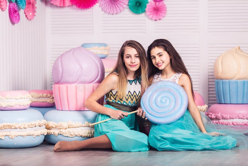 Two beautiful girls in blue dresses hold in their hands huge candy and macaroon in studio with decor of macaroons