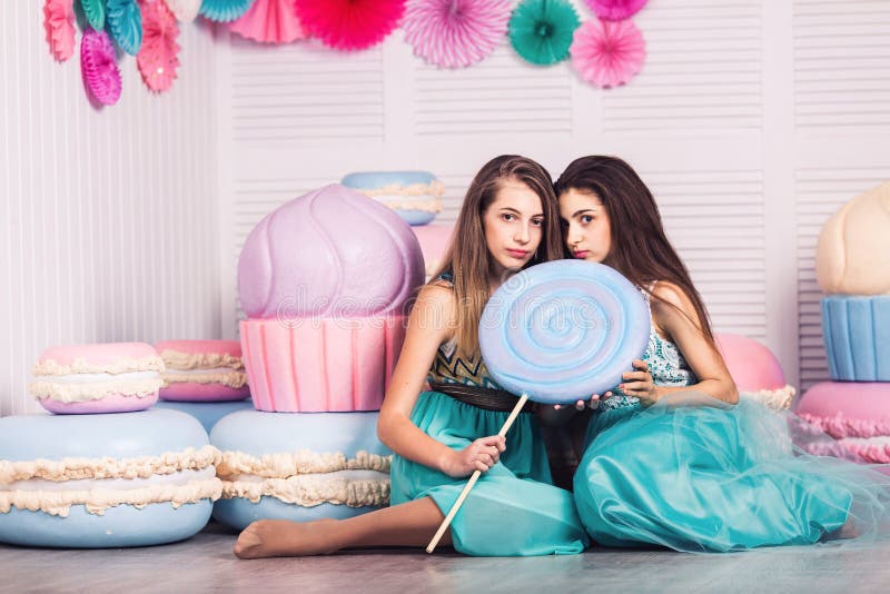 Two beautiful girls in blue dresses hold in their hands huge candy and macaroon in studio with decor of macaroons.