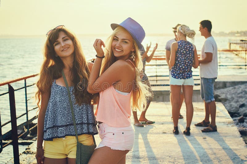 Two beautiful young girls brunette and blonde smiling and having fun at the seaside with group of their friends. Two beautiful young girls brunette and blonde smiling and having fun at the seaside with group of their friends