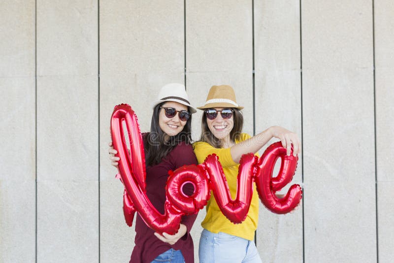 Two beautiful young women having fun outdoors with a red balloon with a love word shape. Casual clothing. They are wearing hats and modern sunglasses. LIfestyle outdoors