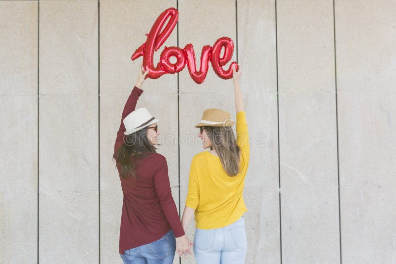 Two beautiful young women having fun outdoors with a red balloon with a love word shape. Casual clothing. They are wearing hats and modern sunglasses. LIfestyle outdoors