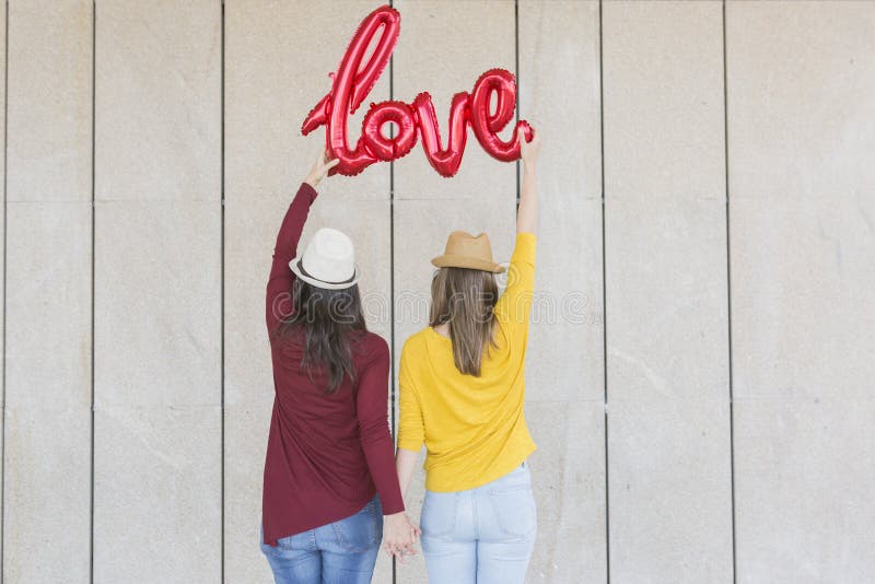 Two beautiful young women having fun outdoors with a red balloon with a love word shape. Casual clothing. They are wearing hats and modern sunglasses. LIfestyle outdoors