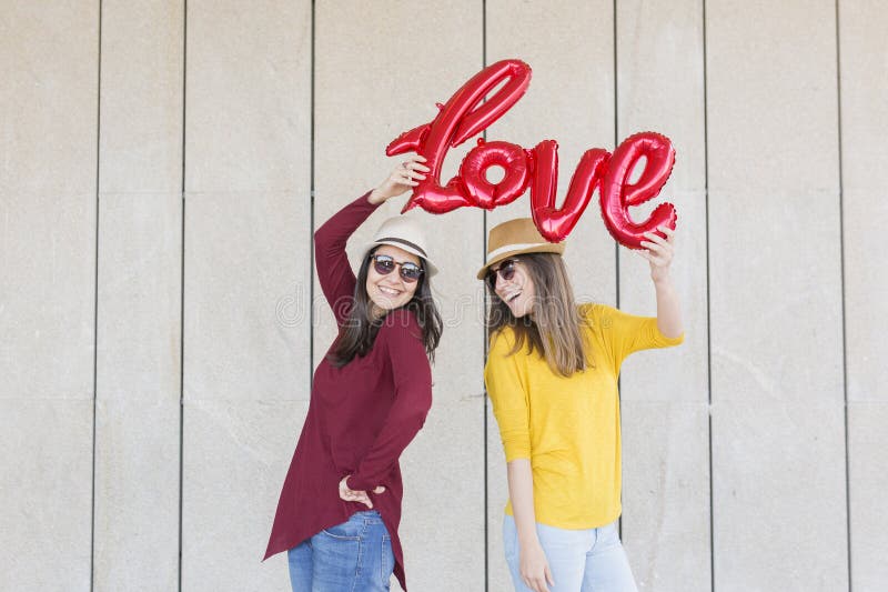 Two beautiful young women having fun outdoors with a red balloon with a love word shape. Casual clothing. They are wearing hats and modern sunglasses. LIfestyle outdoors