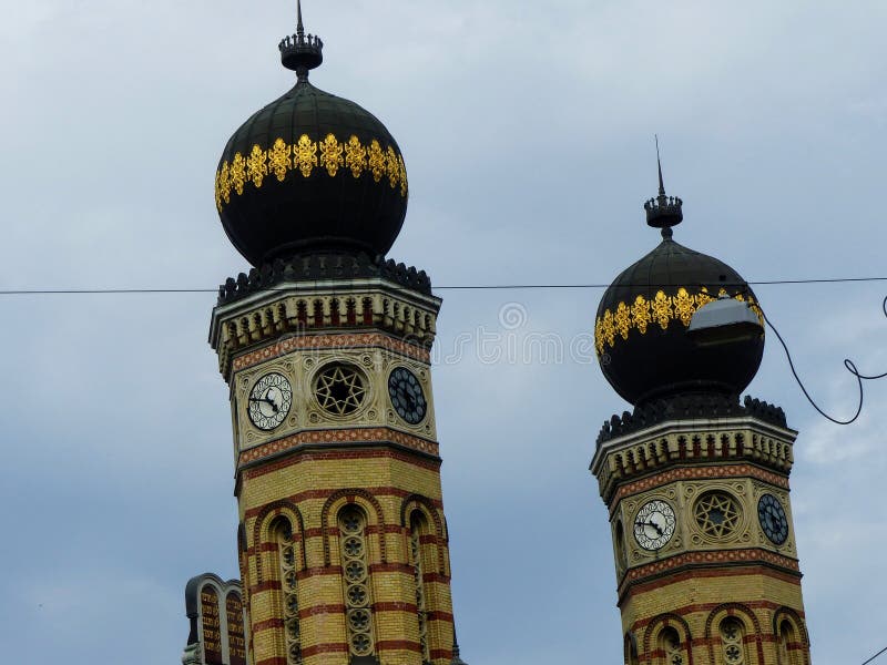 Two bell towers of the synagogue of Budapest in Hungary. Travel destination. Springtime and holidaytime. Blue sky.  Moorish style. Bicolored building. Red stripes. The bigest synagogue of Europe.  Small round decorated windows. Green and golden small domes. Small clocks. Rich decorations. Heritage, culture, art , story and religion.