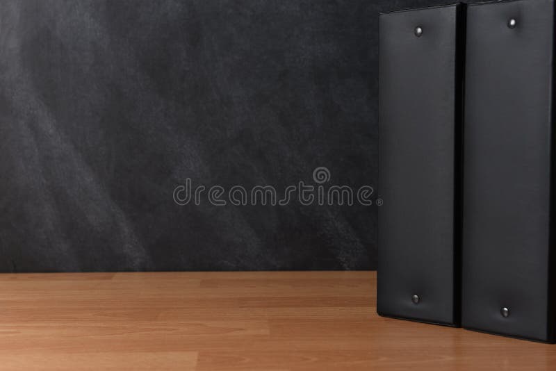 Two black binders standing on a teachers desk in front of a black board