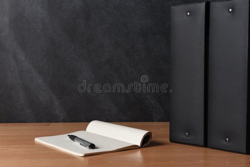 Two black binders standing on a teachers desk with pad and pen in front of a black board