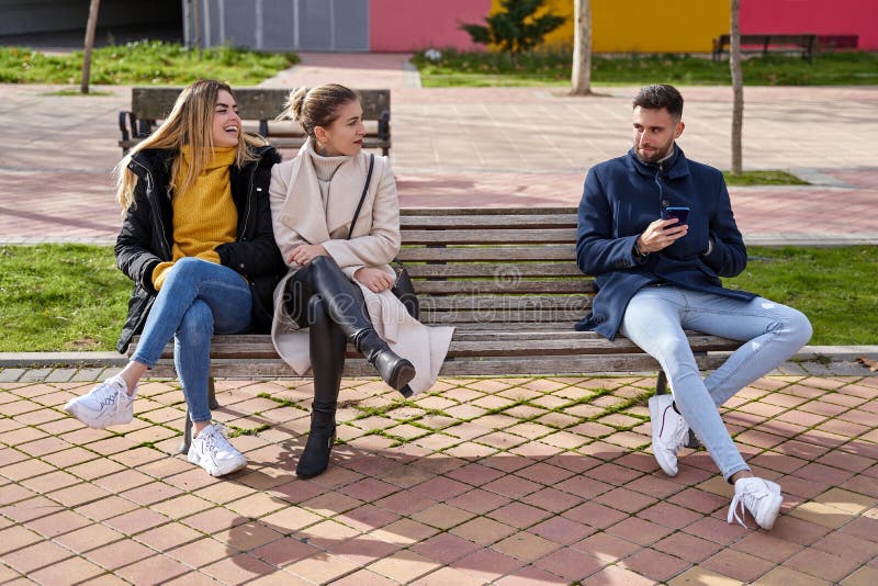 Two blonde girls sitting on a bench next to a guy who is using his cell phone. They are looking at each other