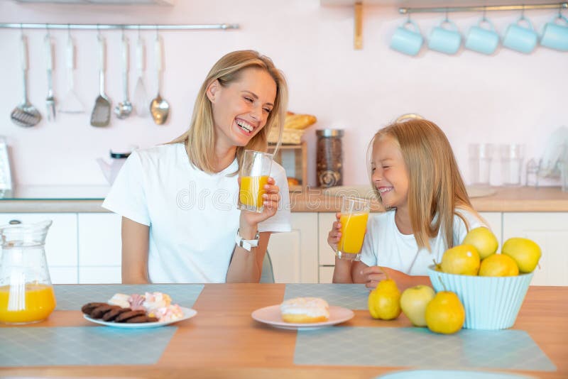 Two blondes, a profile of mom and daughter laugh in the kitchen and drink orange juice. Breakfast with donuts and chocolate chip