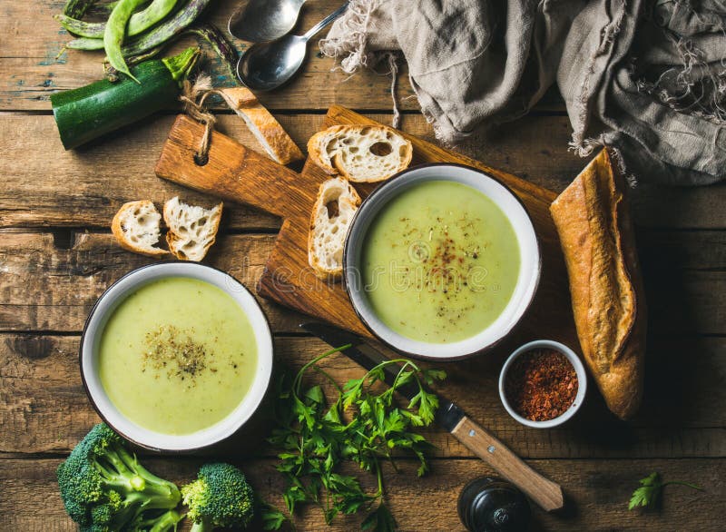 Two bowls of homemade pea, broccoli and zucchini cream soup served with fresh baguette and vegetables on wooden board over rustic background, top view