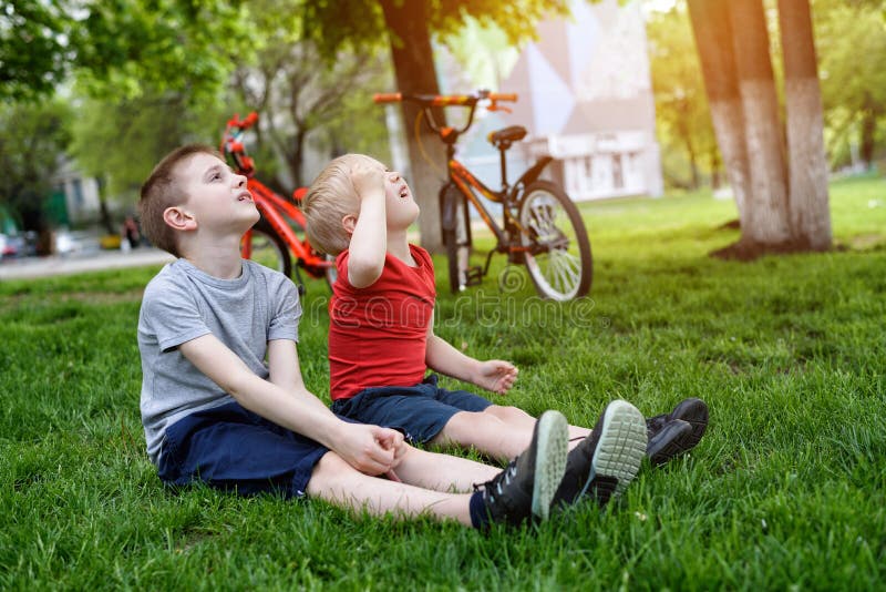 Two boys look up sitting on the grass. Bicycles in the background