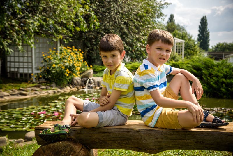 Two brother boys sitting on a bench in a park near peaceful lake with water lily Nymphaeum. Kids relaxing on nature on hot summer