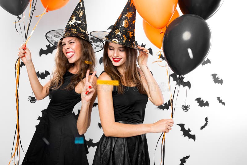 Two brunette girls in black dresses and witch hats have fun with balloons and confetti. Halloween party .