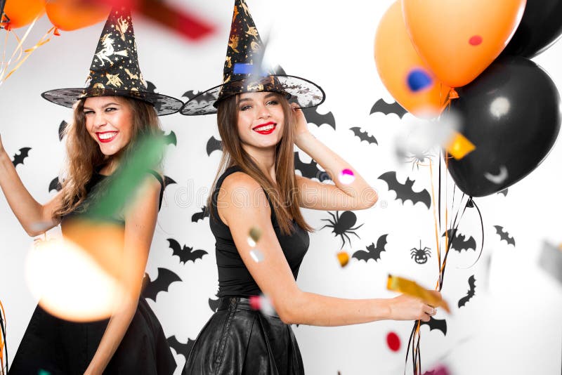 Two brunette girls in black dresses and witch hats have fun with balloons and confetti. Halloween party .