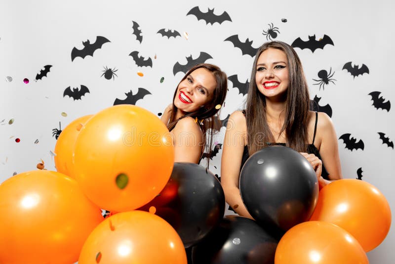 Two brunette women in black dresses smile and hold black and orange balloons. Confetti around. Halloween . .