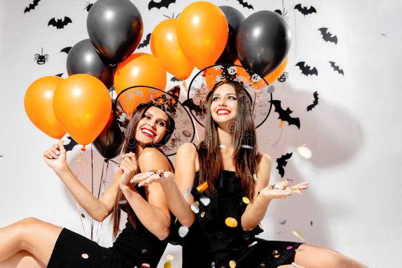 Two brunette women in witches hats smile and hold black and orange balloons. Confetti around. Halloween .