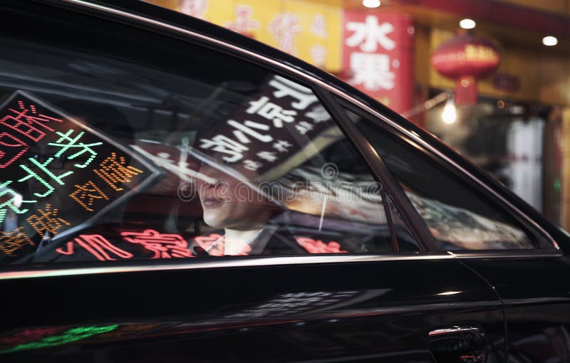 Two business people sitting in the back of a car driving through the city at night, reflections of store signs on the car