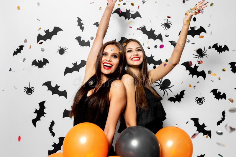 Two charming women in witches hats have fun with confetti and hold black and orange balloons on a white background with black bats and spiders. . .