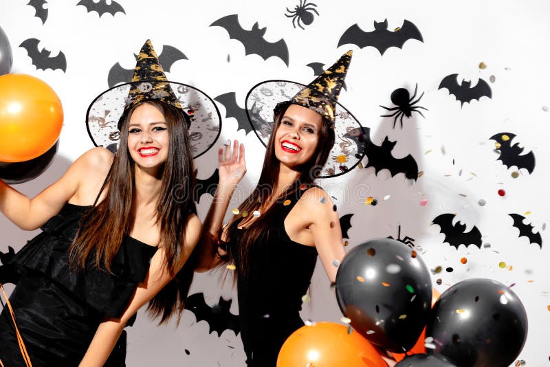 Two charming young women in witches hats hold black and orange balloons on a white background with black bats. Confetti around .