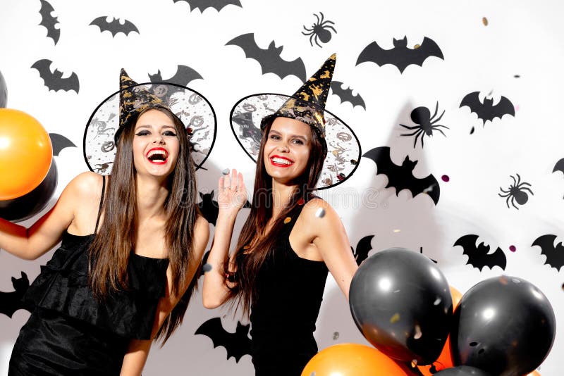 Two charming young women in witches hats hold black and orange balloons on a white background with black bats. Confetti around .