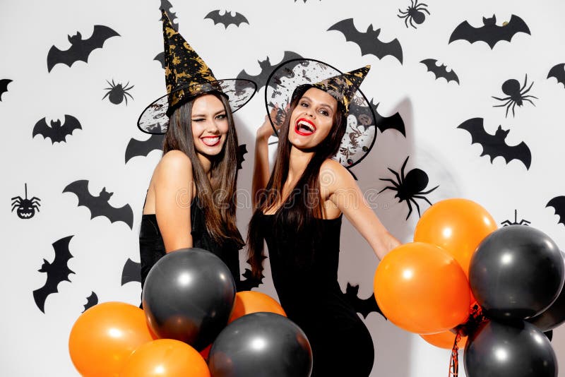 Two charming young women in witches hats hold black and orange balloons on a white background with black bats. Confetti around .