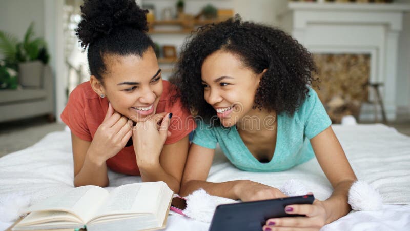 Two cheerful mixed race funny girlfriends reading book and using tablet computer talks and have fun lying in bed at home royalty free stock images Two cheerful mixed race funny girlfriends reading book and using tablet computer talks and have fun lying in bed at home royalty free stock images