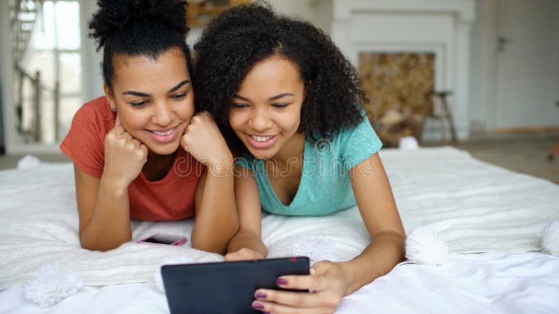 Two cheerful mixed race funny girlfriends sharing social media using tablet computer and talks lying in bed at home royalty free stock photography Two cheerful mixed race funny girlfriends sharing social media using tablet computer and talks lying in bed at home royalty free stock photography