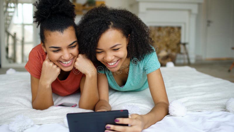 Two cheerful mixed race funny girlfriends sharing social media using tablet computer and talks lying in bed at home stock image Two cheerful mixed race funny girlfriends sharing social media using tablet computer and talks lying in bed at home stock image