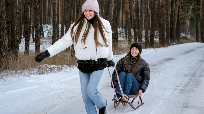 Two cheerful teenage girl riding on sleighs and having fun in winter forest.