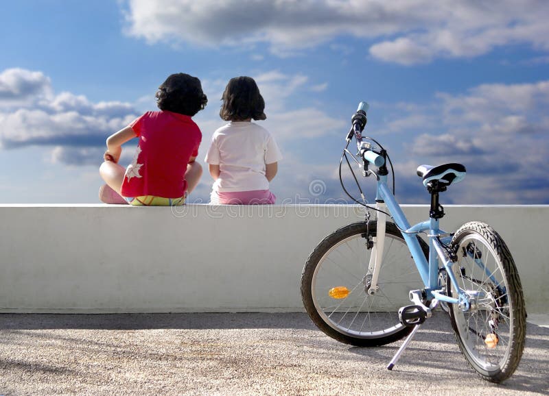 Una bicicletta e due bambini seduti su una parete, guardando verso il cielo, con i loro spalle.