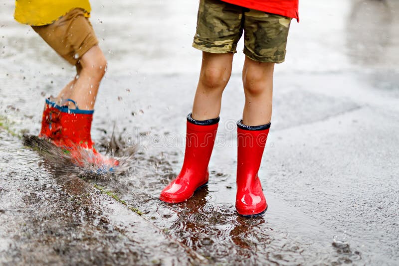 Two children wearing red rain boots jumping into a puddle. Close up. Kids having fun with splashing with water.