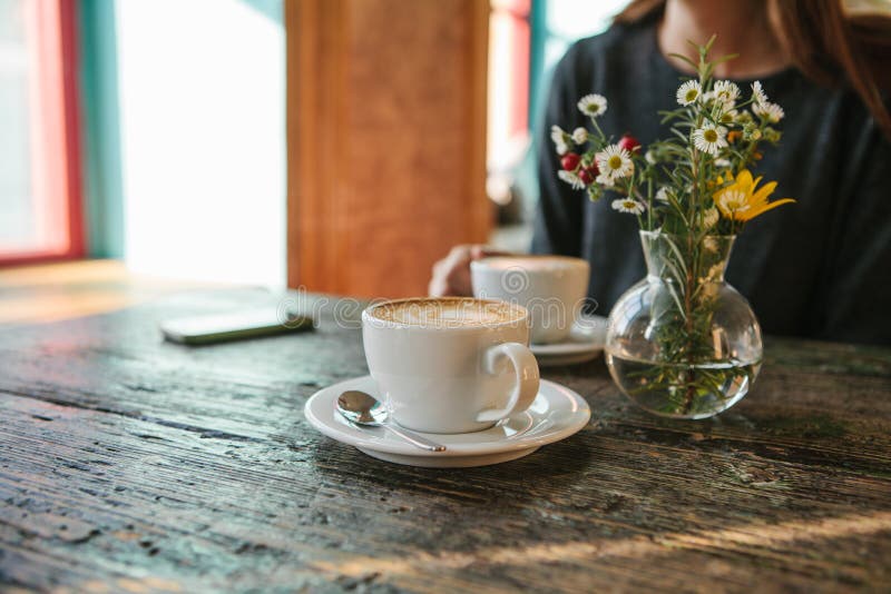 Two cups of coffee and a vase of flowers on a wooden table, the girl holds in her hand one cup of coffee in the